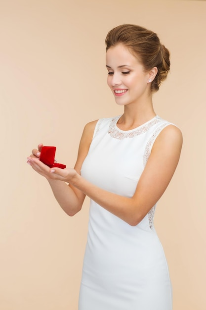 Hochzeits-, Liebes-, Verlobungs- und Glückskonzept - lächelnde Frau in weißem Kleid mit roter Geschenkbox mit Diamantring auf beigem Hintergrund