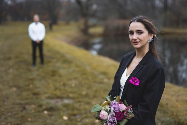 Hochzeitsfotoshooting im Spätherbst Mittleres Porträt einer attraktiven Frau aus dem Nahen Osten Ende 20 in einem Hochzeitskleid und einer Smokingjacke, die in die Kamera blickt Bräutigam Silhouette im Hintergrund