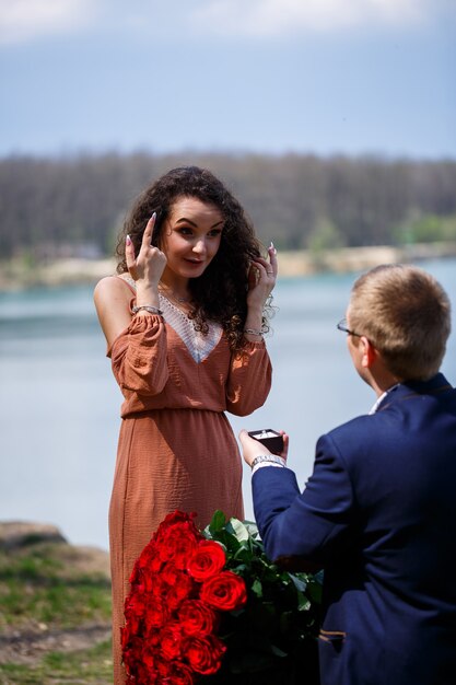 Hochzeitshintergrund im Wald: Ein Mann überrascht, ein Ring gibt einem schönen Mädchen eine Liebeserklärung und einen Heiratsantrag. Mädchen mit einem Lächeln im Gesicht und einem Strauß roter Rosen