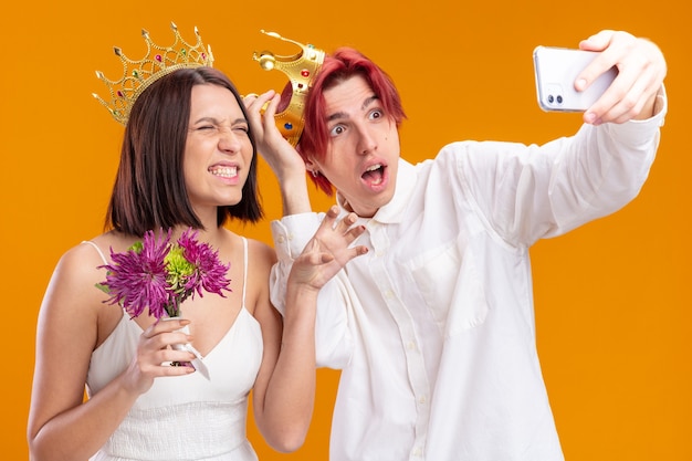 Hochzeitspaar Bräutigam und Braut mit Blumenstrauß im Hochzeitskleid mit goldenen Kronen, die verwirrt und überrascht aussehen, wenn sie Selfie mit dem Smartphone machen