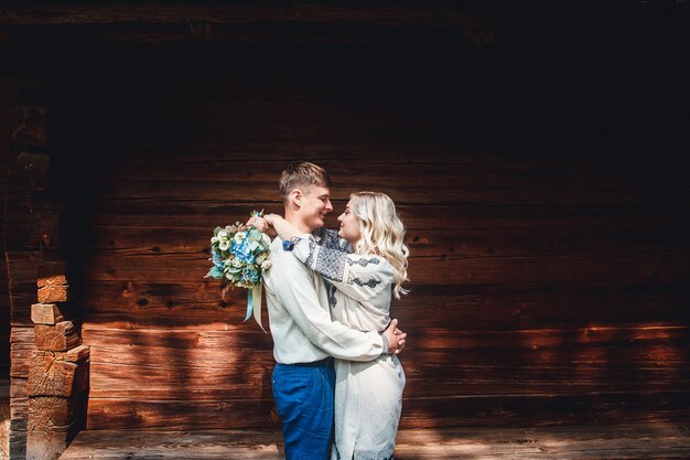 Hochzeitspaar in einem bestickten Hemd mit einem Blumenstrauß auf dem Hintergrund eines Holzhauses