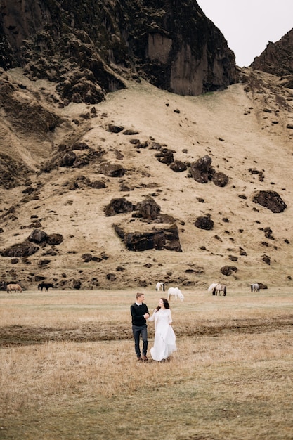 Hochzeitspaar nahe felsigem Berg und grasenden Pferden in Island. Die Braut und