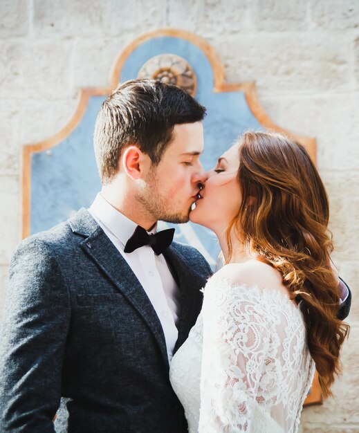 Hochzeitspaarküsse in der Altstadt. Steinmauern der antiken Stadt im Hintergrund. Rustikale Spitzenbraut mit offenem Haar und Bräutigam in grauem Anzug und Fliege. Romantische Liebe in der Straße mit Vintage-Atmosphäre.