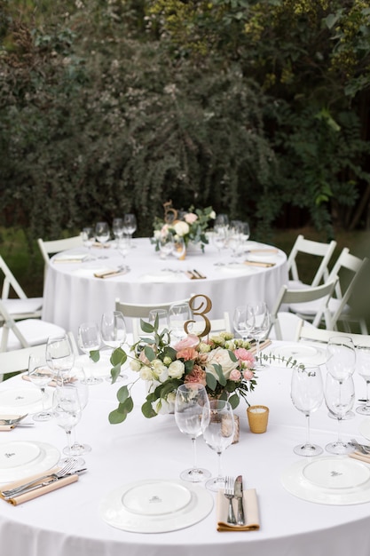 Hochzeitstafeleinstellung verziert mit frischen Blumen in einem Messingvase. Hochzeitsfloristik. Banketttisch für Gäste im Freien mit Blick auf die grüne Natur. Blumenstrauß mit Rosen-, Eustoma- und Eukalyptusblättern.