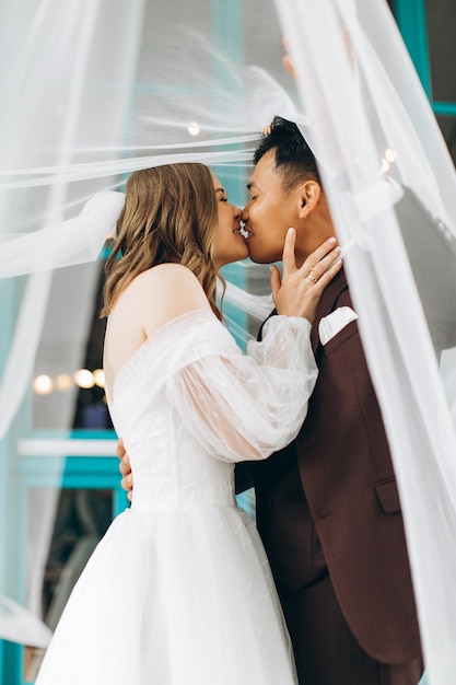 Hochzeitstag Schöne europäische Braut und ihr asiatischer Bräutigam posieren vor dem Hintergrund von Cafés und großen Fenstern