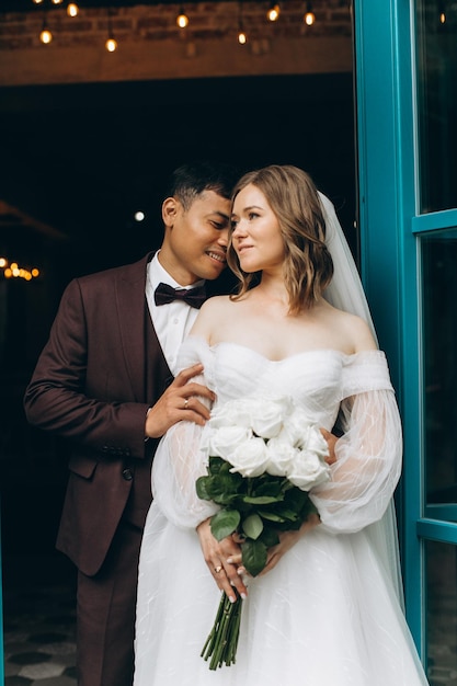 Hochzeitstag Schöne europäische Braut und ihr asiatischer Bräutigam posieren vor dem Hintergrund von Cafés und großen Fenstern