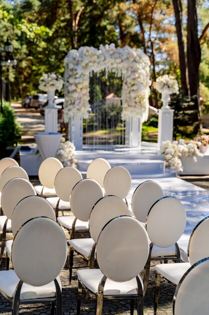 Hochzeitstorbogen mit Blumen im Park für eine Hochzeitszeremonie angeordnet. Zarter Bogen zum Feiern. Raws von Stühlen auf Vorderansicht.