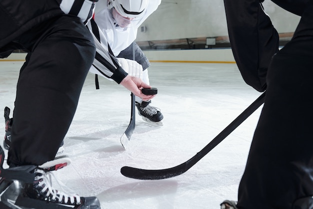 Hockey-Schiedsrichter hält Puck über Eisbahn, während zwei Rivalen mit Stöcken sich nach vorne beugen und auf den Moment warten, um der erste zu sein, um ihn zu schießen