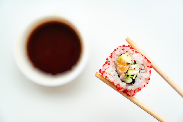 Hölzerne Sushi-Sticks halten Rollen mit rotem Kaviar, Fisch, Reis und Avocado mit einer Tasse Sojasauce auf weißem Hintergrund. Essen mit Lieferung online bestellen. Japanische Küche.