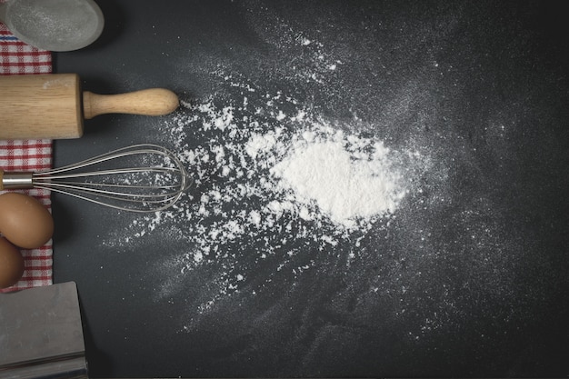 Hölzerne Teigrolle und Schneebesen auf dem schwarzen Tisch mit Mehlaufstrich