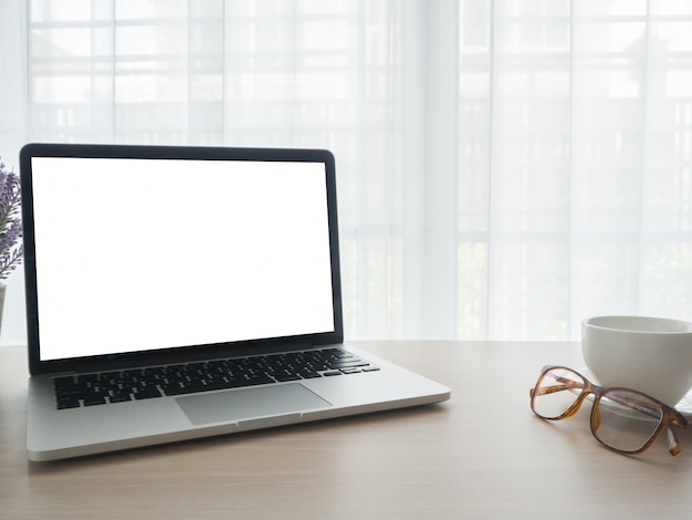 Hölzerner Bürotisch mit Laptop des leeren Bildschirms auf weißer Kaffeetasse auf Vorhangfenster backgrou