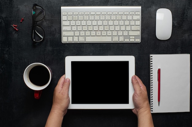 Hölzerner schwarzer Schreibtischtisch mit der Hand, die einen digitalen leeren Bildschirmtisch mit Notebook-Brillen und anderen Vorräten mit Draufsicht einer Tasse Kaffee hält