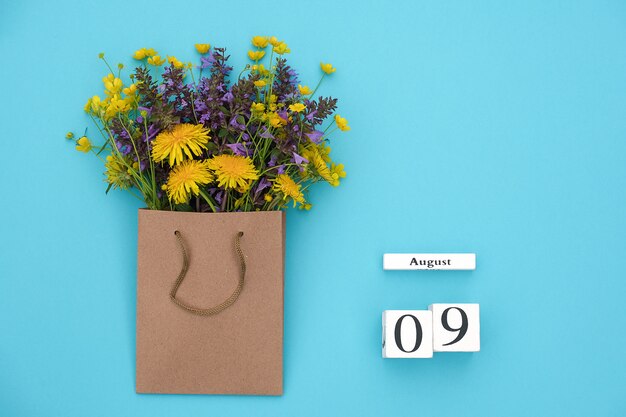 Hölzerner würfelkalender am 9. august und bunte rustikale blumen des feldes im handwerkspaket auf blau.