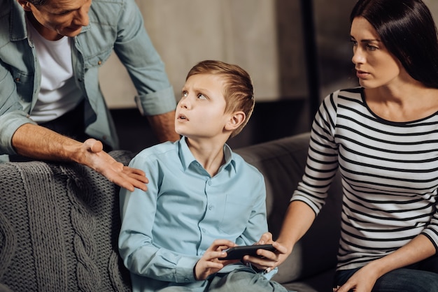 Hör auf. Strenger junger Vater schimpft mit seinem Sohn, weil er am Telefon Binge-Playing-Spiele spielt, während der Junge ihn empört ansieht