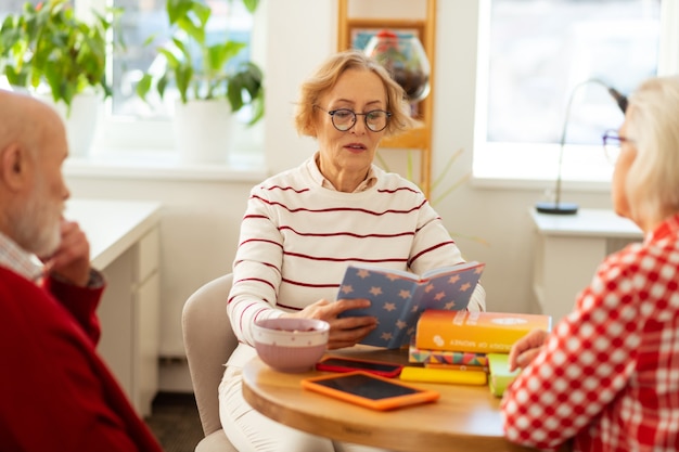 Hör zu. Gut aussehende, angenehme Frau, die in das Buch schaut, während sie es ihren Freunden vorliest