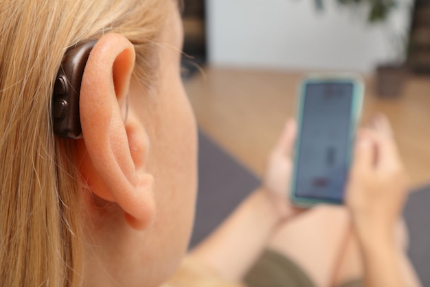 Hörbehinderte Frau arbeitet mit einem Smartphone, Nahaufnahme einer blonden Frau, die ein Hörgerät am Ohr trägt und einen Knopf drückt, während sie zu Hause ein Smartphone benutzt