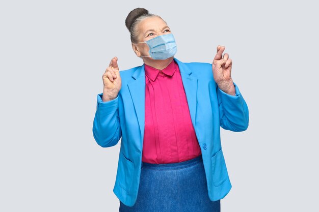 Hoffnungsvoll mit chirurgischer medizinischer Maske kreuzte Frau die Finger und wünschte. Großmutter mit hellblauem Anzug und rosa Hemd stehend mit gesammelten Brötchenhaaren. Indoor-Studioaufnahme, isoliert auf grauem Hintergrund