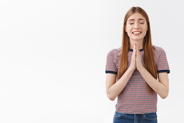 Hoffnungsvolle, positive, süße europäische Frau in gestreiftem T-Shirt hält die Hände im Gebet und lächelt nervös und optimistisch, die Augen schließen, glauben, dass Träume wahr werden und Gott bitten, den Wunsch zu erfüllen, weißer Hintergrund