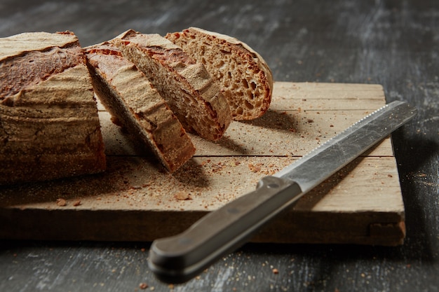 Hogaza de pan integral en rodajas sobre una placa de pan de madera con un cuchillo, sobre un fondo oscuro