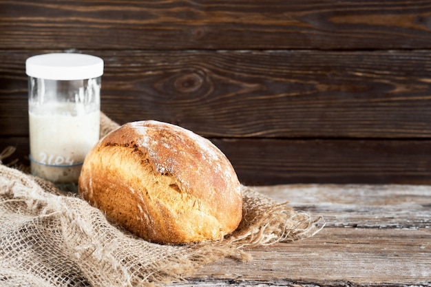 Hogaza de pan de masa madre de leche blanca casera, enfoque selectivo. un vaso de masa madre sobre la mesa. primer plano, con espacio de copia. pan artesanal sobre un forro de tela, fondo de madera