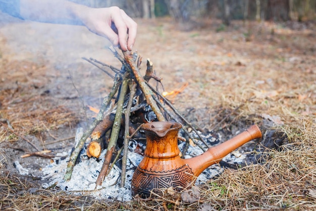 En la hoguera en el bosque de primavera, una cafetera turca de arcilla se calienta contra la hierba En el marco, la mano de un hombre