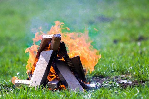 Hoguera que brilla intensamente en la naturaleza. Quema de tablones de madera afuera el día de verano. Brillantes llamas anaranjadas, humo ligero y cenizas oscuras sobre la hierba verde en verde borrosa. Concepto de turismo y camping.