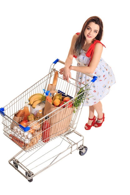 Hohe Betrachtungswinkel eines Mädchens, das in die Kamera lächelt, während es einen Einkaufswagen voll mit Lebensmitteln auf weißem Hintergrund schiebt.