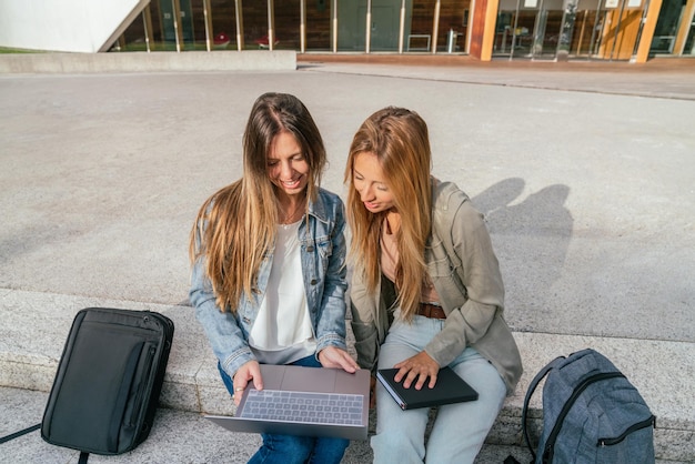 Hohe Betrachtungswinkel von zwei jungen Studentinnen, die an einem sonnigen Tag mit einem Laptop auf der Universitätstreppe sitzen.