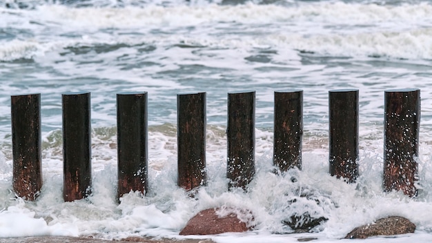 Hohe hölzerne Wellenbrecher in blau spritzenden Meereswellen, Nahaufnahme. Lange Stangen oder Buhnen im Wasser. Stürmischer Tag auf See.