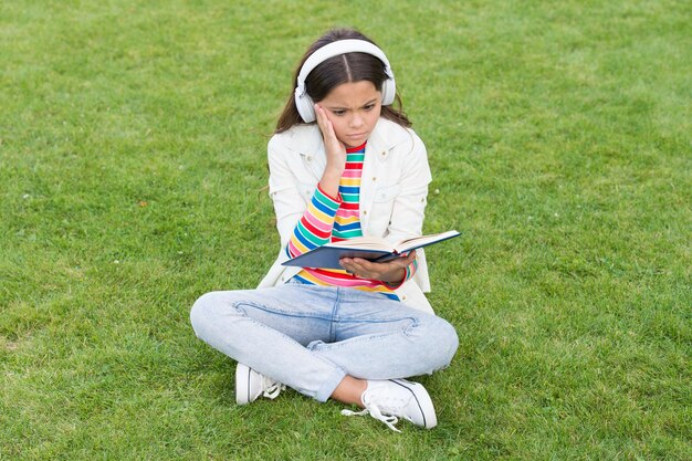 Hohe Standards erreichen Kleines Mädchen, das Buchliteratur für Kinder liest, schreibt Kindheitserinnerungen, intelligentes Kind im Headset, zurück zur Schule, Kind lernt online im Park, hört Musik auf grünem Gras mit Buch