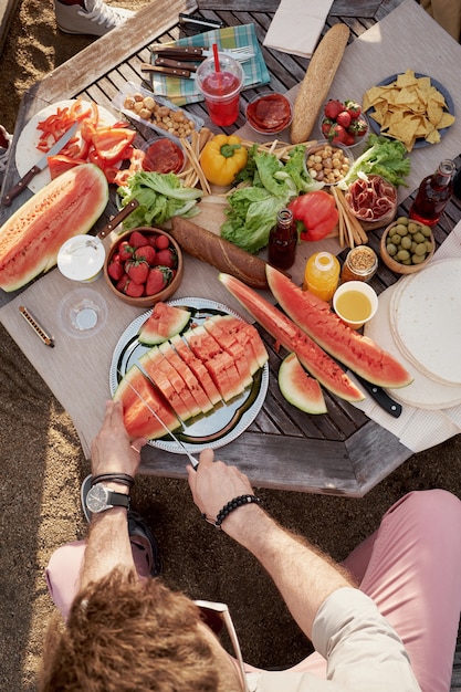 Hohe Winkelansicht des nicht erkennbaren Mannes, der reife Wassermelone im Freien beim Sitzen am Tisch mit appetitlichem frischem Obst und Gemüse schneidet