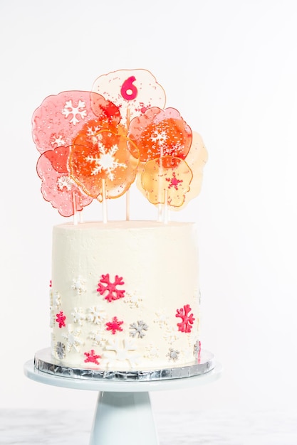 Hoher runder Kuchen mit italienischem Buttercreme-Zuckerguss, verziert mit Fondant-Schneeflocken und gekrönt mit großen rosa und weißen Lutschern auf weißem Hintergrund.