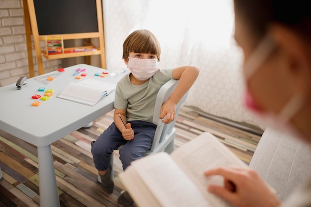 Hoher Winkel des Lehrers mit der medizinischen Maske, die Kind zu Hause unterrichtet