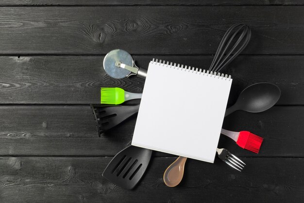 Hoja en blanco de bloc de notas abierto y utensilios de cocina en la mesa con mantel, espacio de copia