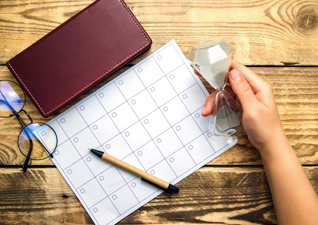 Foto una hoja en blanco de bloc de notas y un calendario para planificar un mes o un año. gafas, bloc de notas y bolígrafo sobre un fondo de madera. espacio de trabajo la mano de una mujer sostiene un reloj de arena.