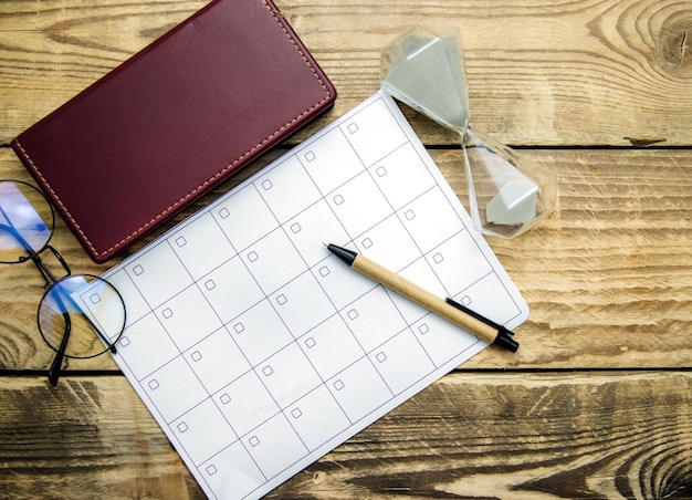 Una hoja en blanco de bloc de notas y un calendario para planificar un mes o un año. Gafas, reloj de arena, bloc de notas y bolígrafo sobre un fondo de madera. espacio de trabajo