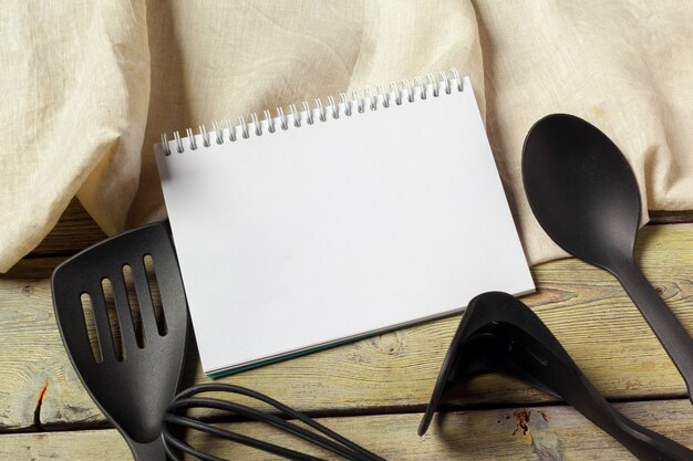 Hoja en blanco de libreta abierta y utensilios de cocina en la mesa con mantel, espacio de copia