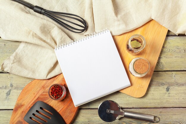 Hoja en blanco de libreta abierta y utensilios de cocina en la mesa con mantel, espacio de copia