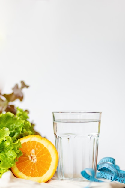 Hoja de ensalada. Naranja, taza de agua y cinta métrica azul sobre fondo blanco con espacio para copiar texto, estilo de vida saludable y concepto de agua potable