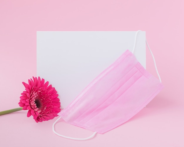 Hoja de papel en blanco con máscara protectora médica y flor de gerbera rosa sobre fondo rosa