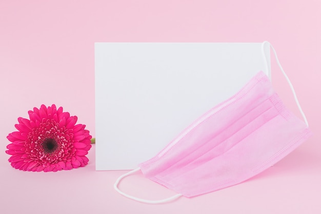 Hoja de papel en blanco con máscara protectora médica y flor de gerbera rosa