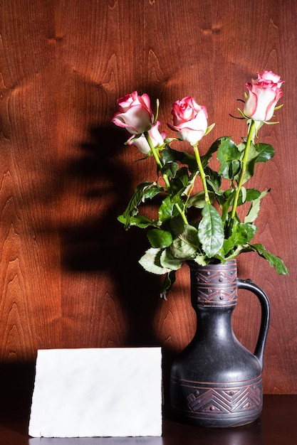 Hoja de papel en blanco y rosas rosadas en jarra de cerámica