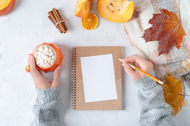 Hoja de papel en blanco sobre cuaderno con manos de mujer en suéter y taza de calabazas.