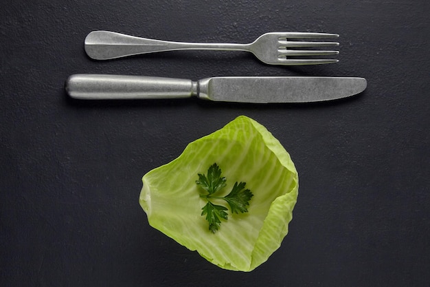 Una hoja de repollo blanco con una hoja de perejil y un tenedor con un cuchillo sobre un fondo de hormigón negro Concepto de nutrición vegetariana para la salud y la pérdida de peso