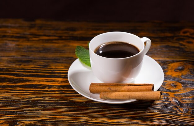 Hoja con té negro en taza y plato junto a un par de ramas de canela sobre mesa de madera