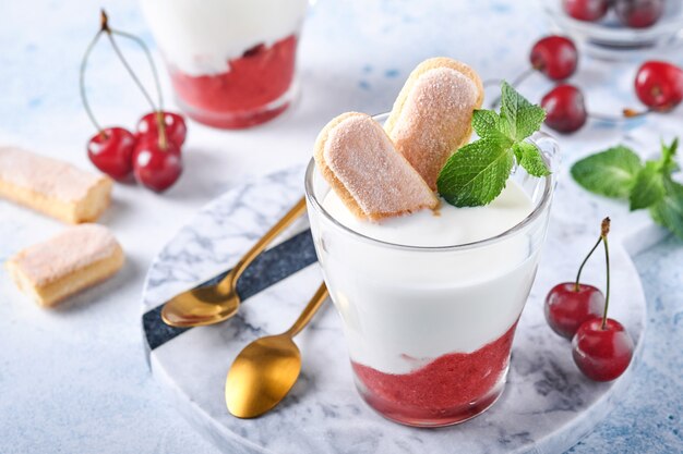 Hojaldre de cereza de verano con galletas savoiardi y queso crema en vidrio sobre fondo gris claro. Pastel de tiramisú tradicional con frutos rojos frescos. Enfoque selectivo.