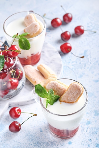 Hojaldre de cereza de verano con galletas savoiardi y queso crema en vidrio sobre fondo gris claro. Pastel de tiramisú tradicional con frutos rojos frescos. Enfoque selectivo.