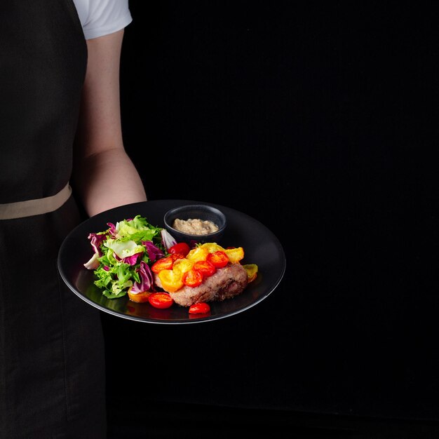 Hojas de ensalada de bistec a la parrilla servidas en un plato negro sobre una presentación de fondo negro en manos del chef