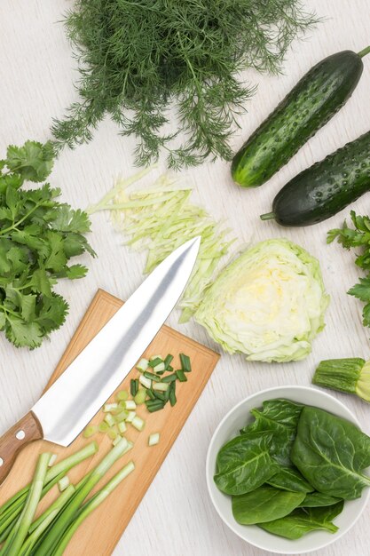 Hojas de espinaca en un tazón blanco. Cebollas verdes y cuchillo sobre tabla de cortar. Repollo, pepinos y verduras en la mesa. Fondo blanco. Vista superior