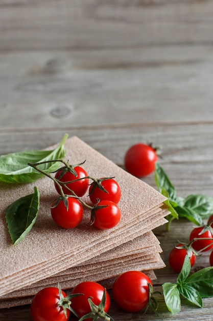 Hojas de lasaña cruda, albahaca y tomates cherry en una mesa de madera